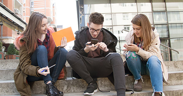 Three smiling teenagers sit on a step outside as they talk and tap their phones