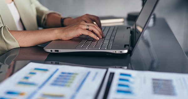 Blog on optimizing college student retention: Female administrator typing on a laptop with printouts of graphs on her desk